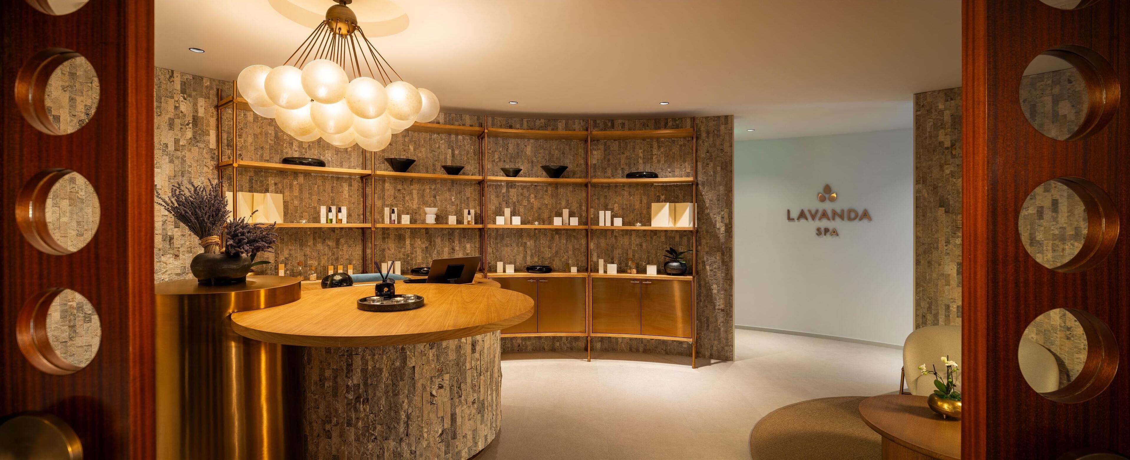A modern reception area with a central wooden counter, shelves with various items, and a chandelier.
