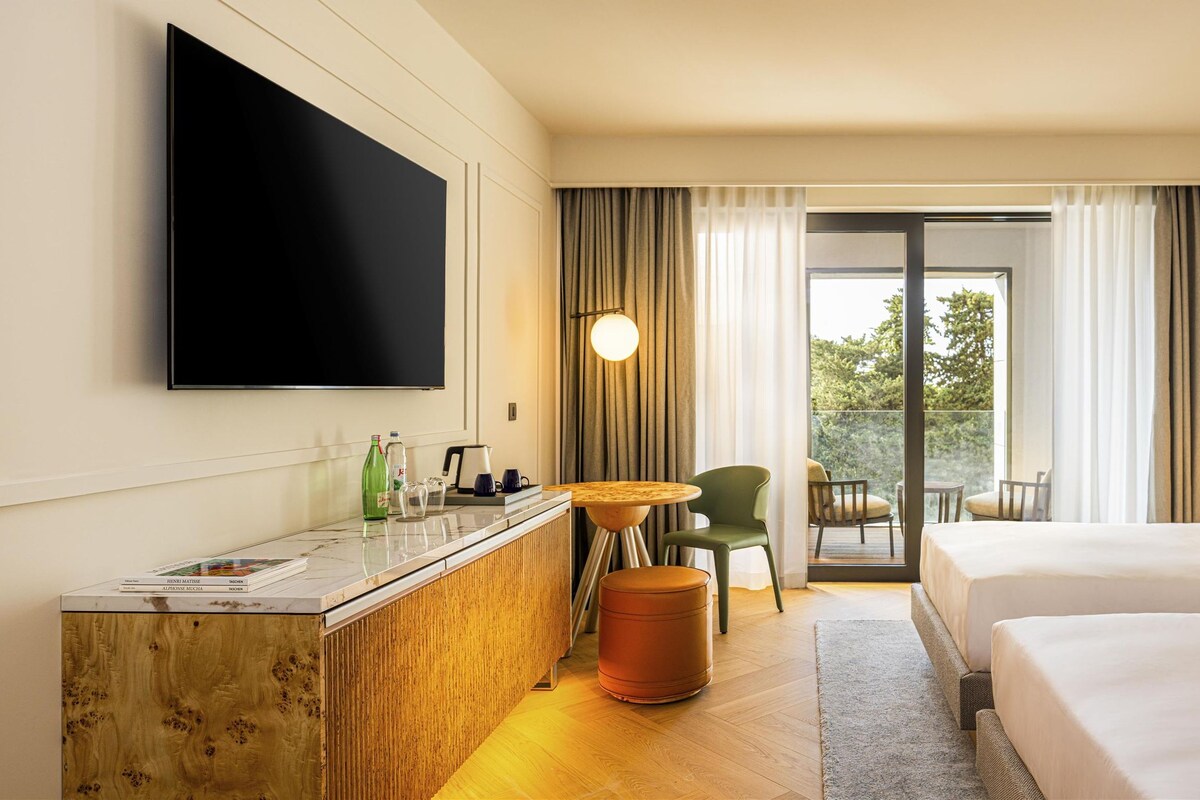 Ein Hotelzimmer mit zwei Betten, einem Schreibtisch, einem Flachbildfernseher, einem kleinen Tisch und Blick ins Grüne durch das Fenster.