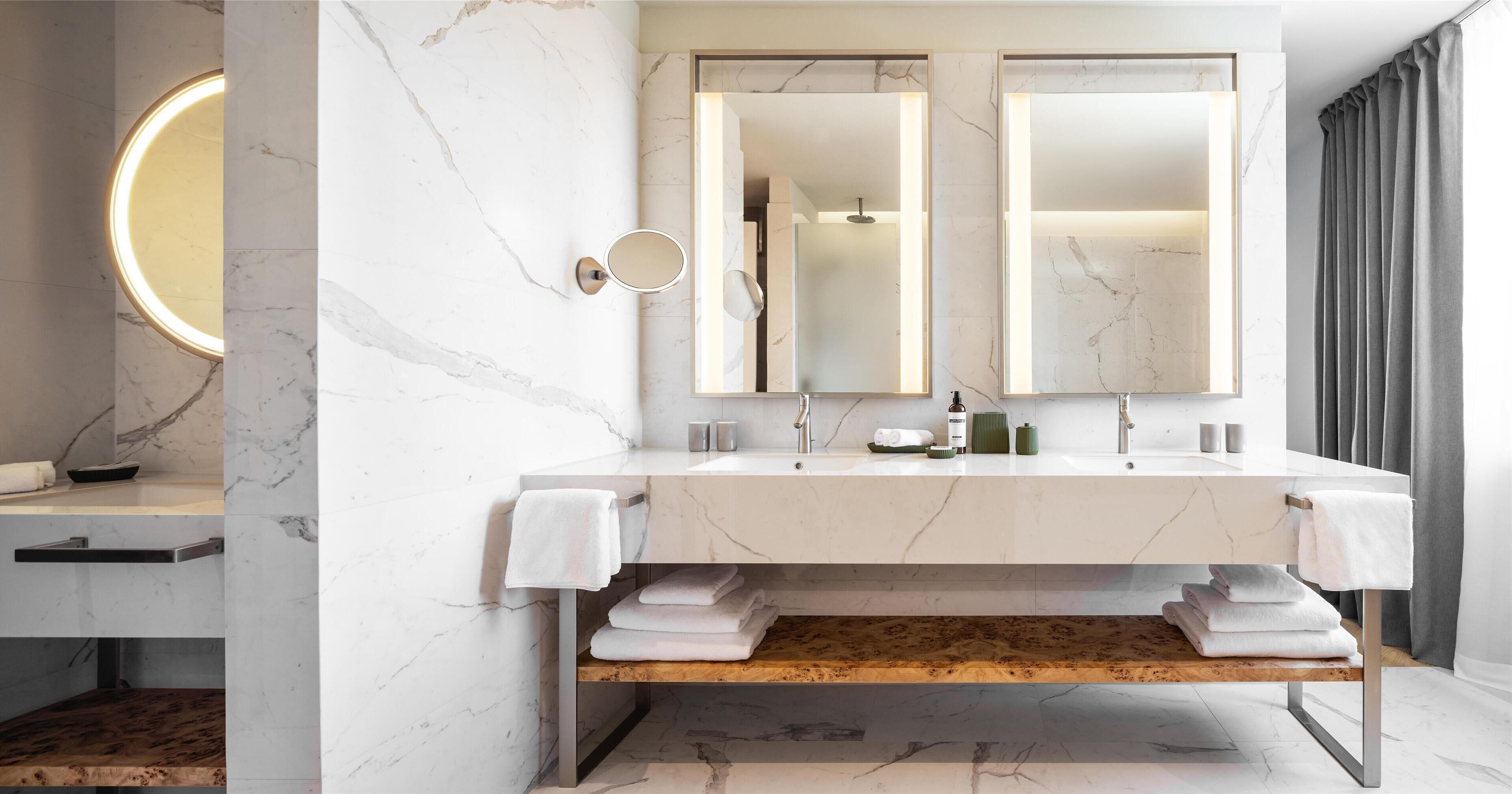 A modern bathroom with a marble vanity, two mirrors, a towel rack, and a wooden shelf.