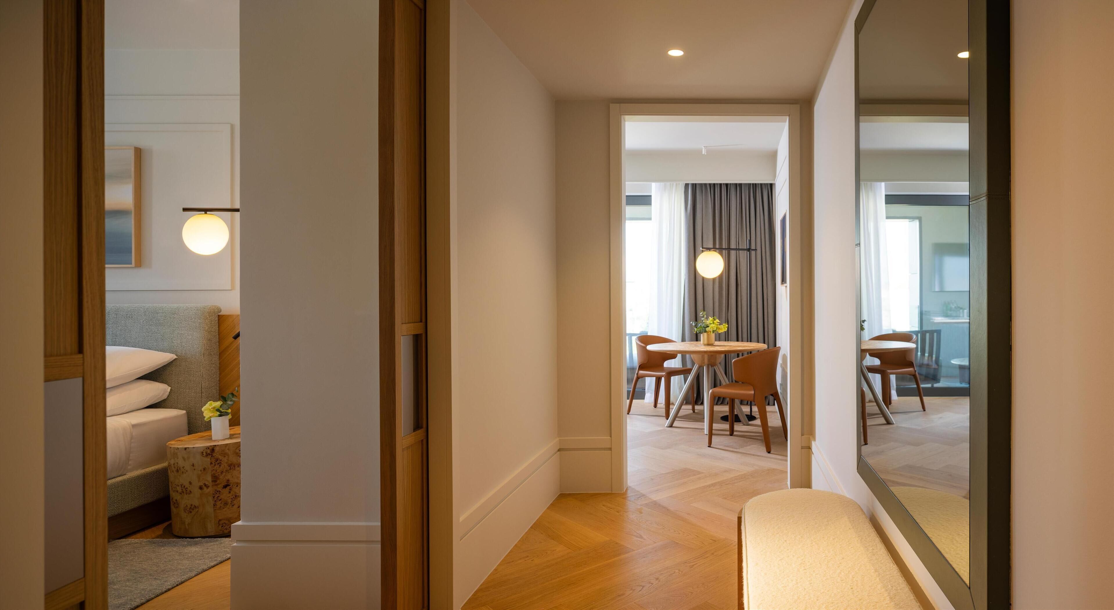 A modern hotel room with a wooden floor, a bed with white linens, a bedside table with a lamp, and a view of the dining area through a glass door.