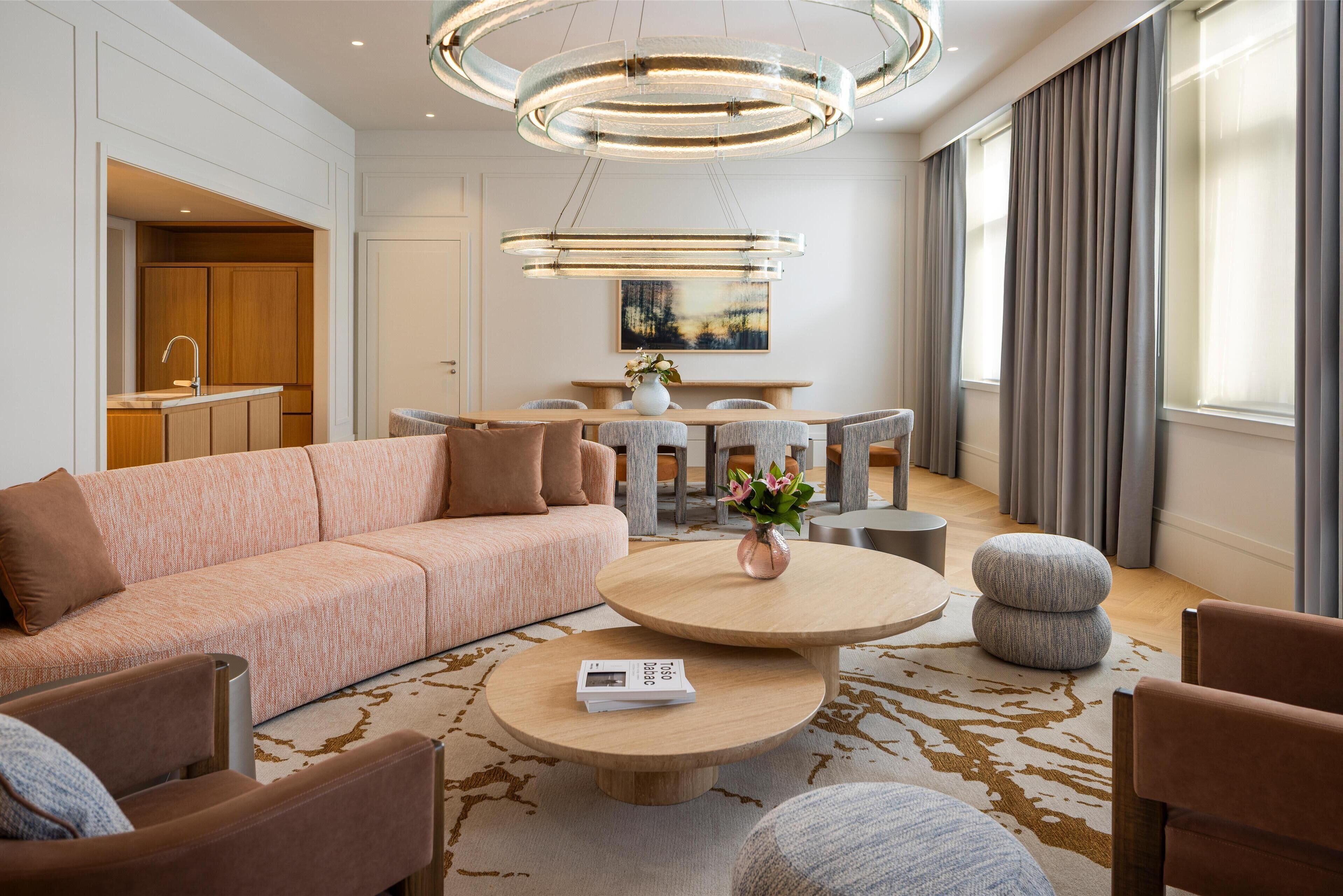 A modern living room with a sofa, coffee table, and ottomans. There is a dining area with a table and chairs, and a kitchen in the background.