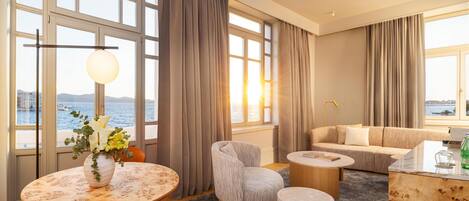 Ein modernes Hotelzimmer mit großen Fenstern, einem runden Tisch mit einer Vase, einem Sofa, einem Couchtisch und einem Holznachttisch.