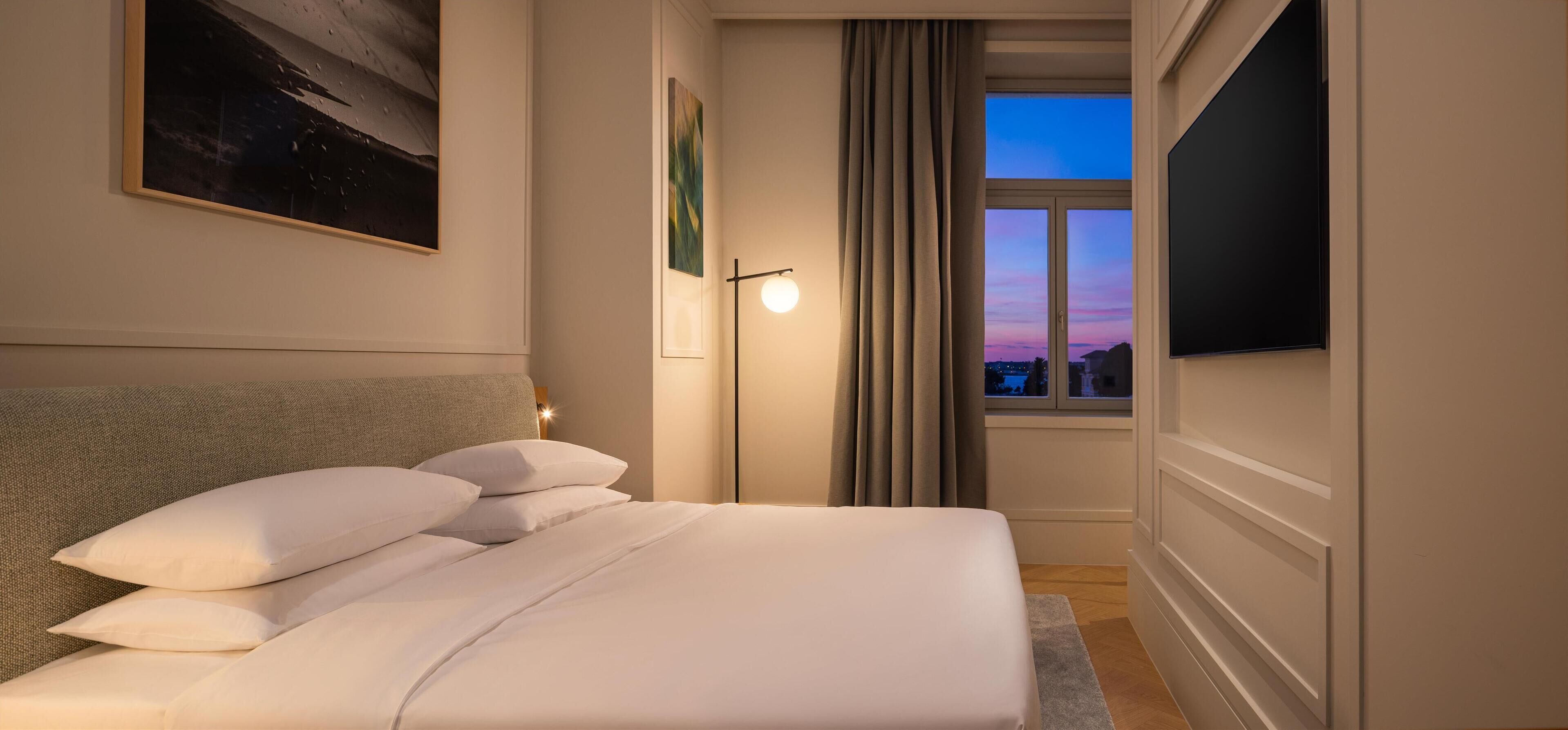A neatly made bed with pillows, a bedside lamp, a painting on the wall, and a window with curtains.