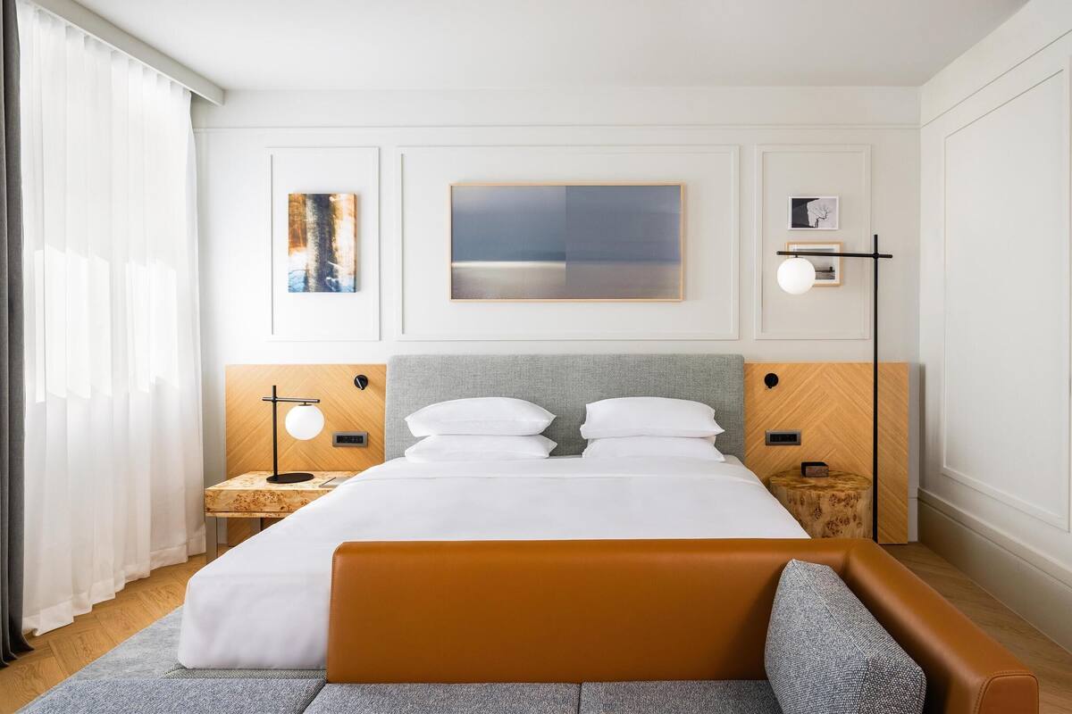 A modern hotel room with a large bed, bedside tables, a nightstand, a sofa, a bathroom with a marble sink, and artwork on the walls.