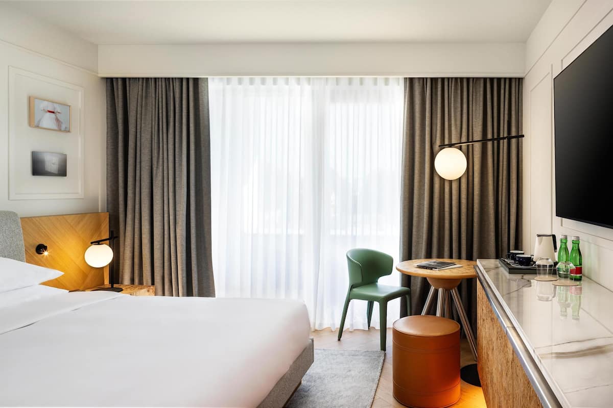 A hotel room with a bed, a desk with a lamp, a chair, a round table with a tray of items, and a TV.