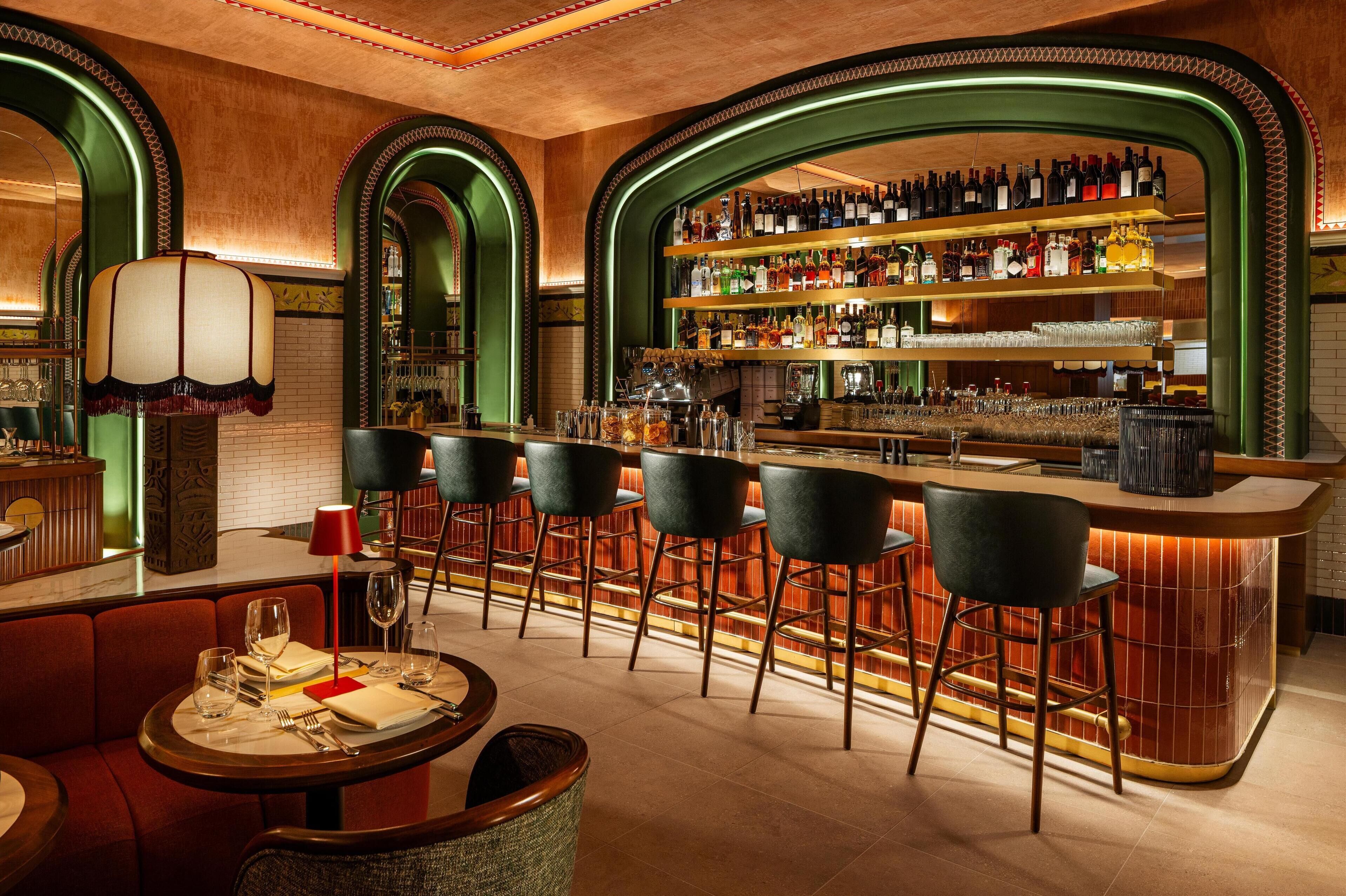 A bar area with a long counter, multiple stools, and a variety of bottles and glasses on shelves.