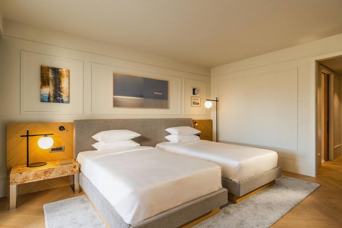 A hotel room with two single beds, a bedside table with a lamp, a framed picture on the wall, and a door to the right.