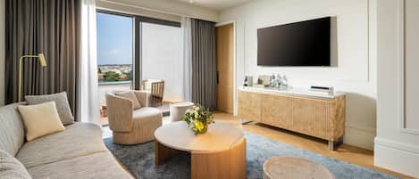 A modern hotel room with a large sofa, a round coffee table, a TV mounted on the wall, and a view of the city through large windows.