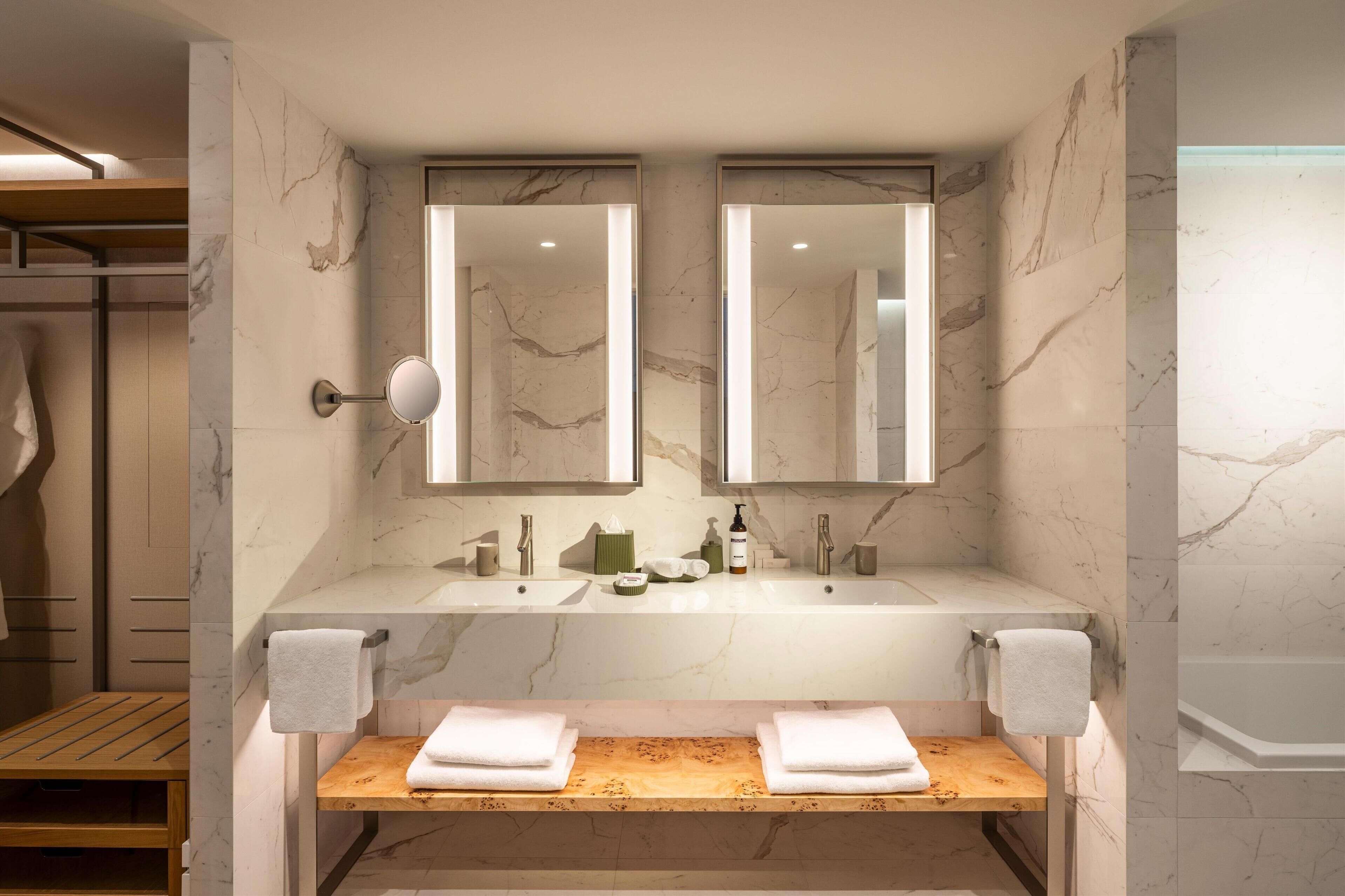 A modern bathroom with two sinks, marble walls, and a wooden bench.