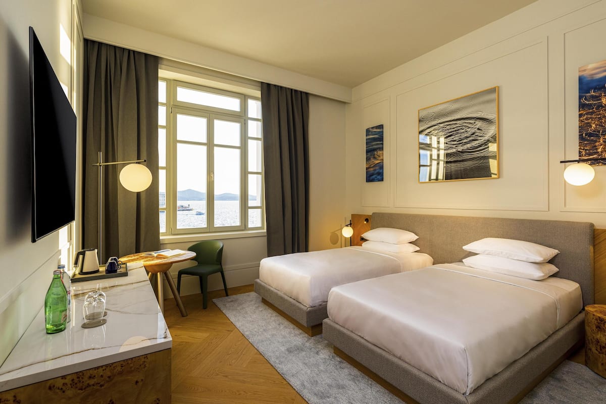 A hotel room with two beds, a desk with a chair, a TV, and a window with curtains.