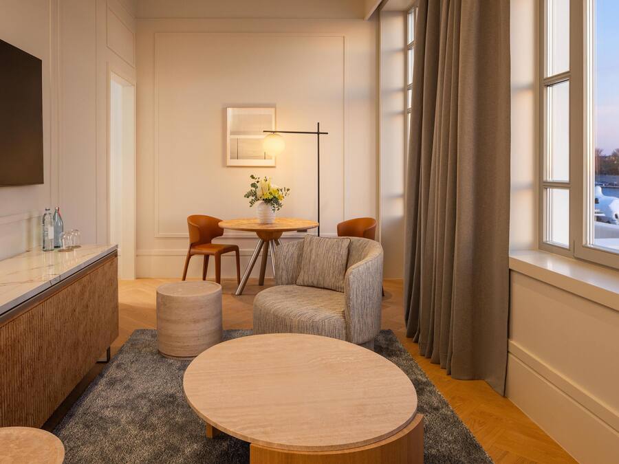 Un salon moderne avec une table basse ronde en bois, deux fauteuils gris à revêtement, une petite table ronde ornée d’un vase de fleurs, un lampadaire et une grande fenêtre avec des rideaux.