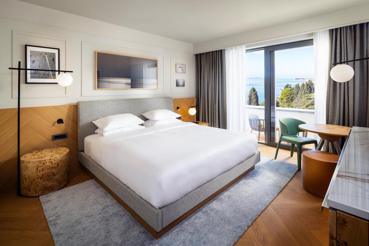 A modern hotel room with a large bed, a dining area, and a balcony with a view of the sea.