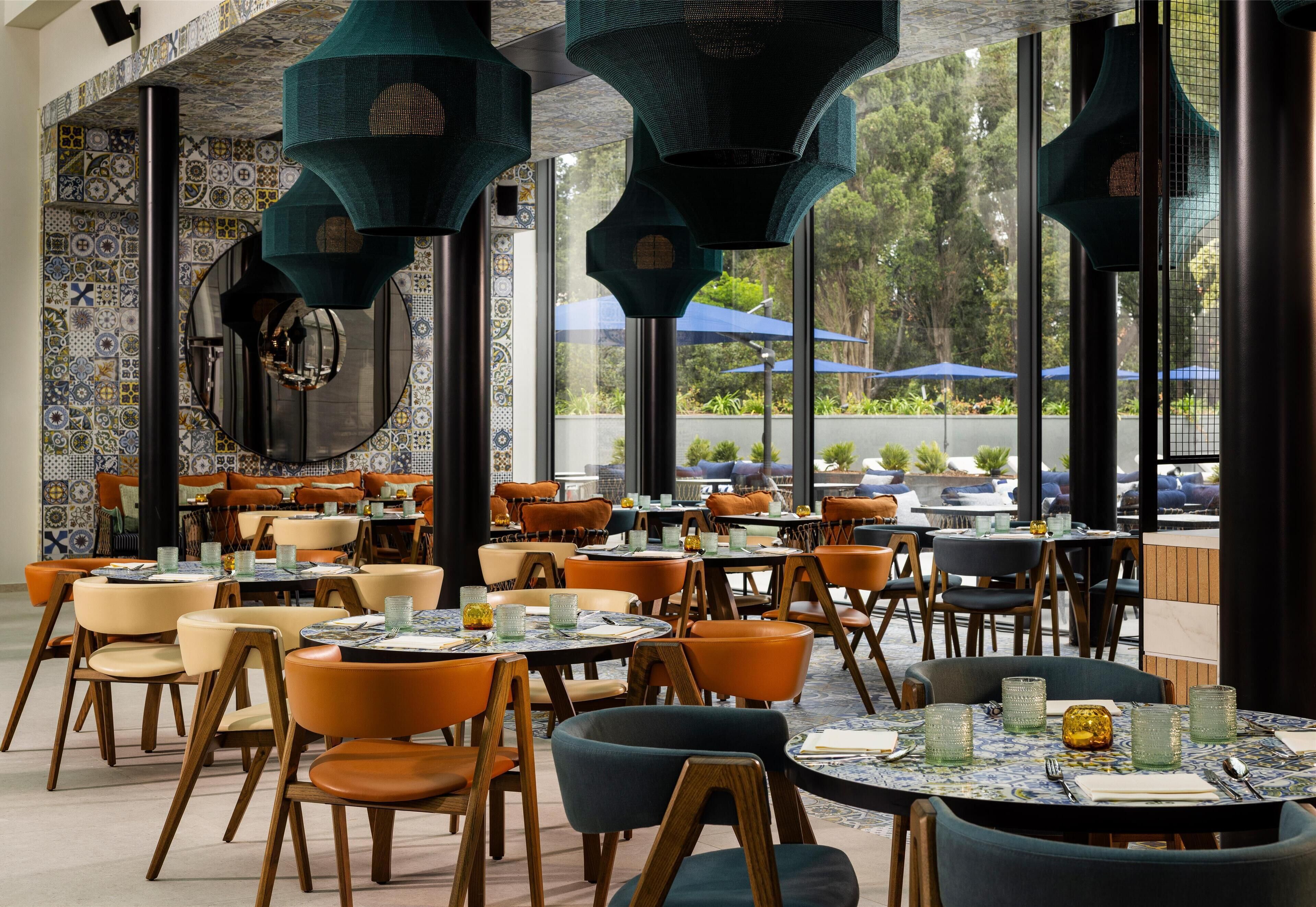 Een restaurant met ronde tafels en oranje stoelen, grote ramen en hangende groene lampen.