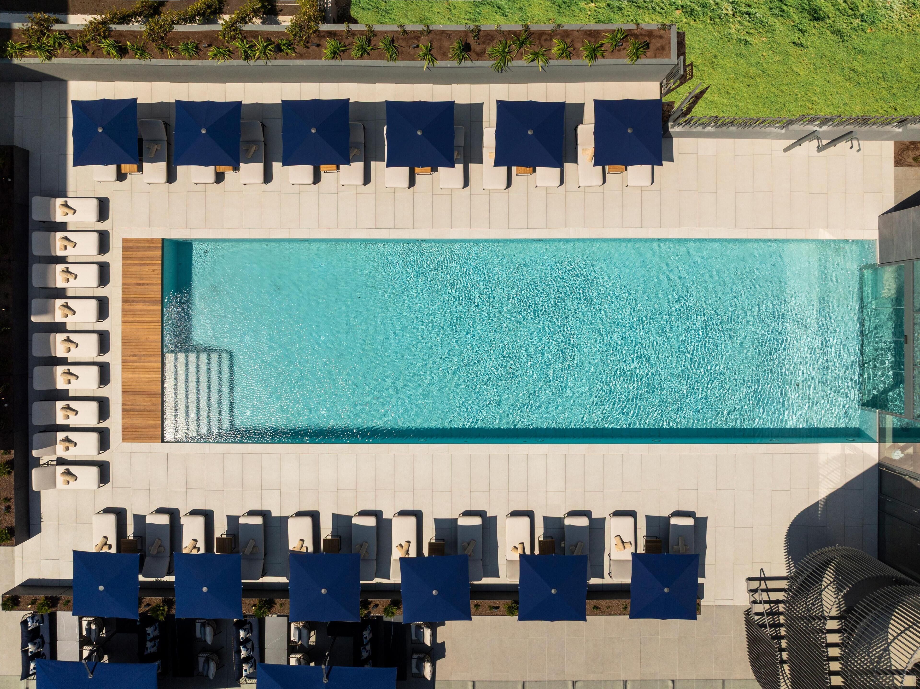 Vista aérea de una piscina rectangular con varias sillas de descanso y sombrillas azules dispuestas a su alrededor.