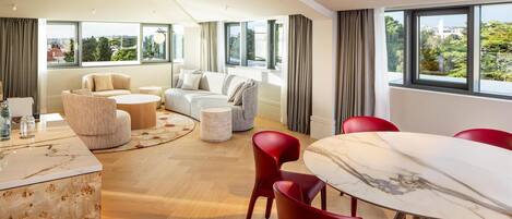A modern hotel room with a large window, a sofa set, a dining table, and red chairs.