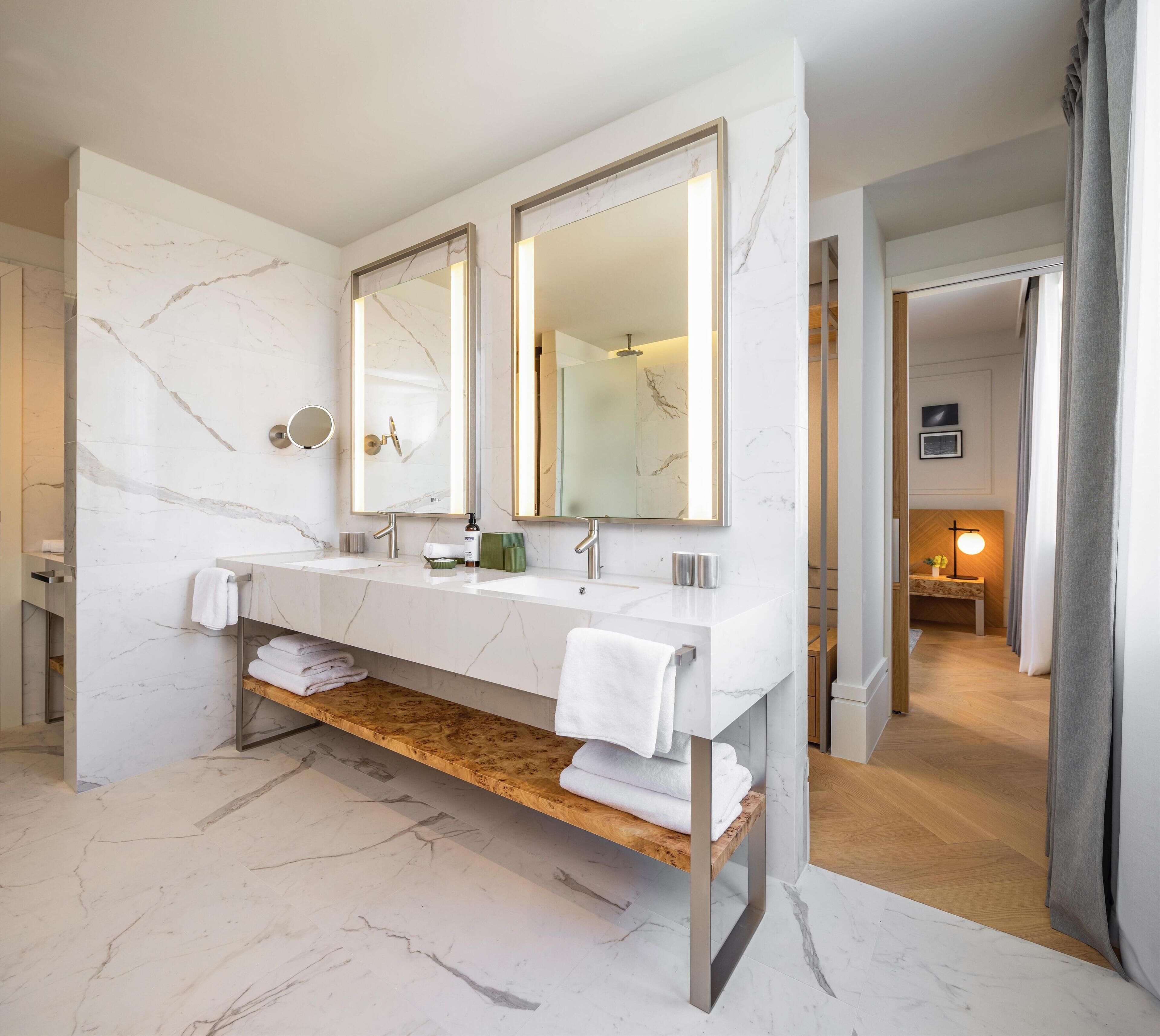 Une salle de bain moderne avec un meuble-lavabo en marbre, deux vasques, un grand miroir et une étagère en bois où sont posées des serviettes.