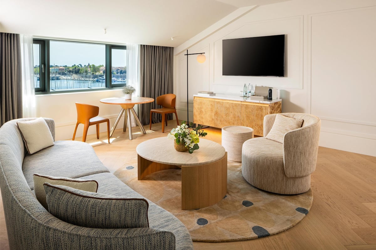 A modern living room with a sofa, armchairs, a coffee table, and a dining area with chairs. A flat-screen TV is mounted on the wall, and a large window offers a view of the marina.