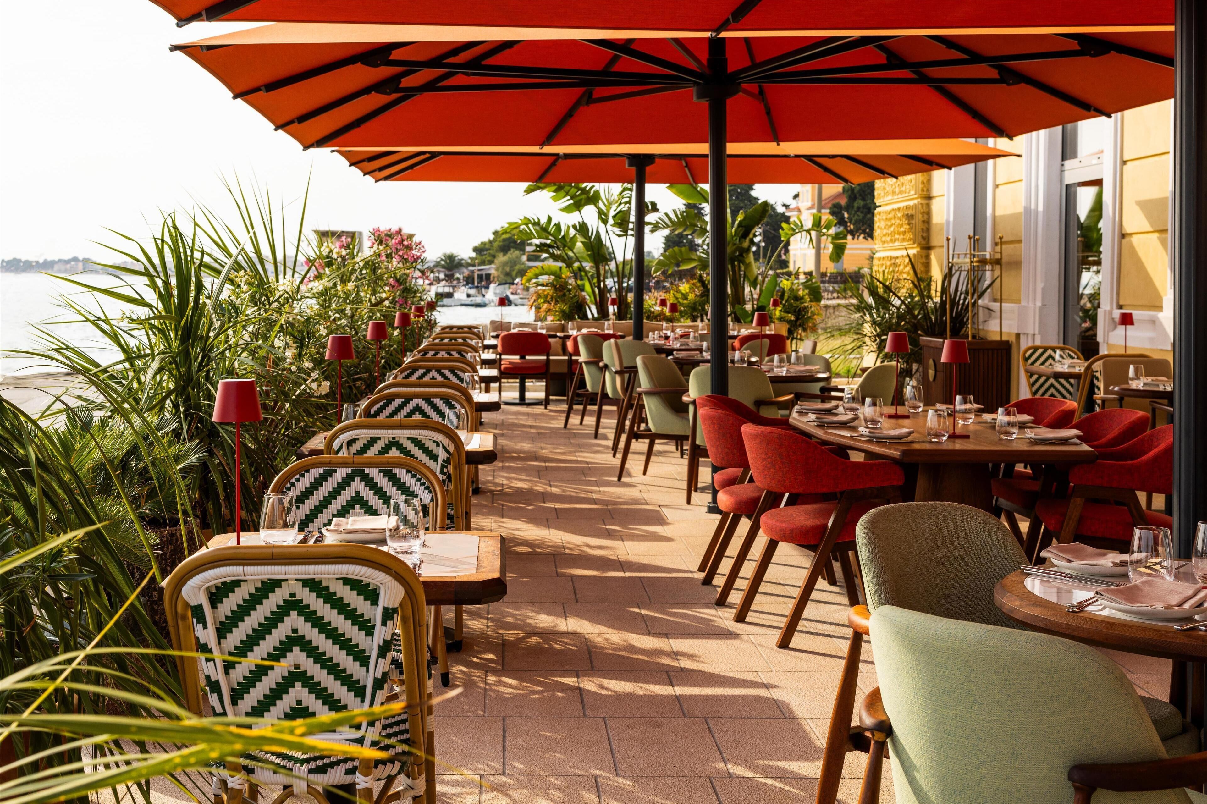 Un espacio de comedor al aire libre con mesas, sillas, sombrillas y plantas en macetas.