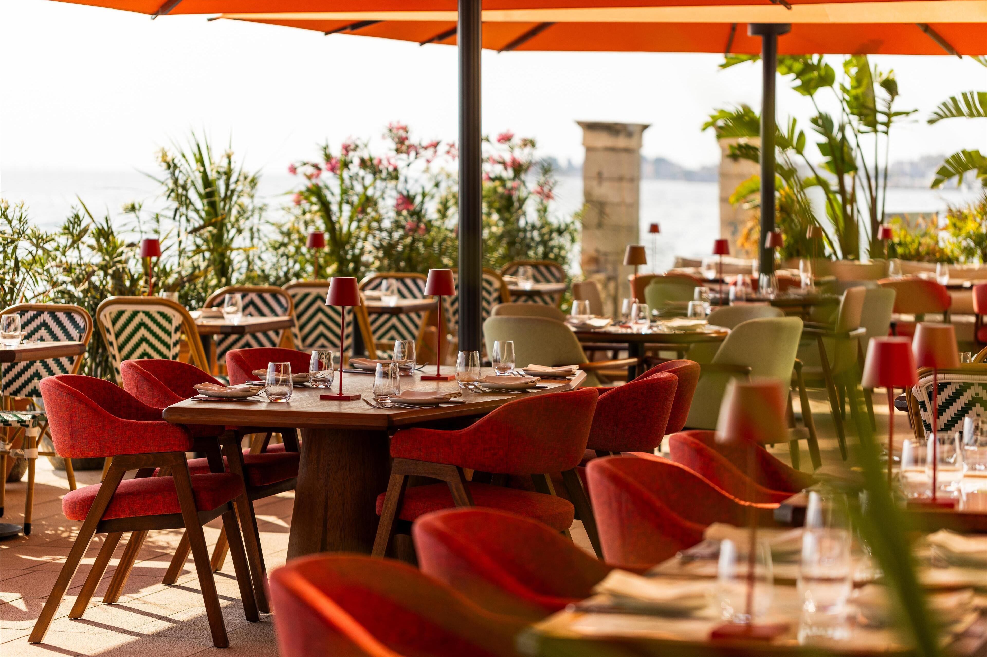 An outdoor dining area with tables and chairs, some with umbrellas, and a view of the sea.