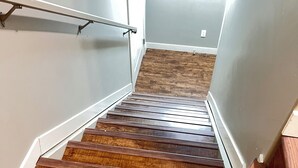 Un escalier en bois avec une rampe métallique, menant à une pièce dont le mur est blanc et le plancher en bois.