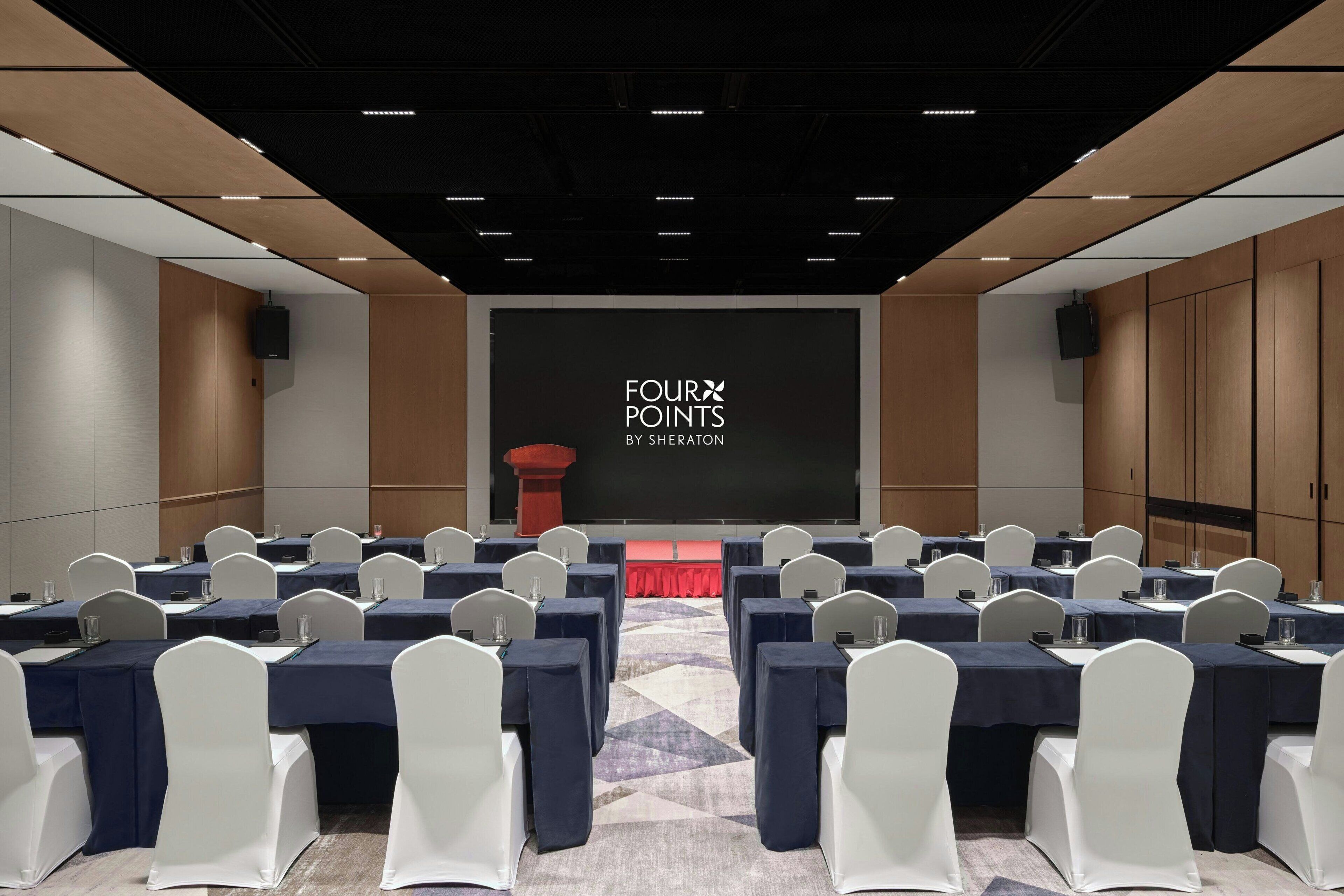 A conference room with rows of tables and chairs, a stage with a screen displaying "FOUR POINTS BY SHERATON", and speakers on the walls.