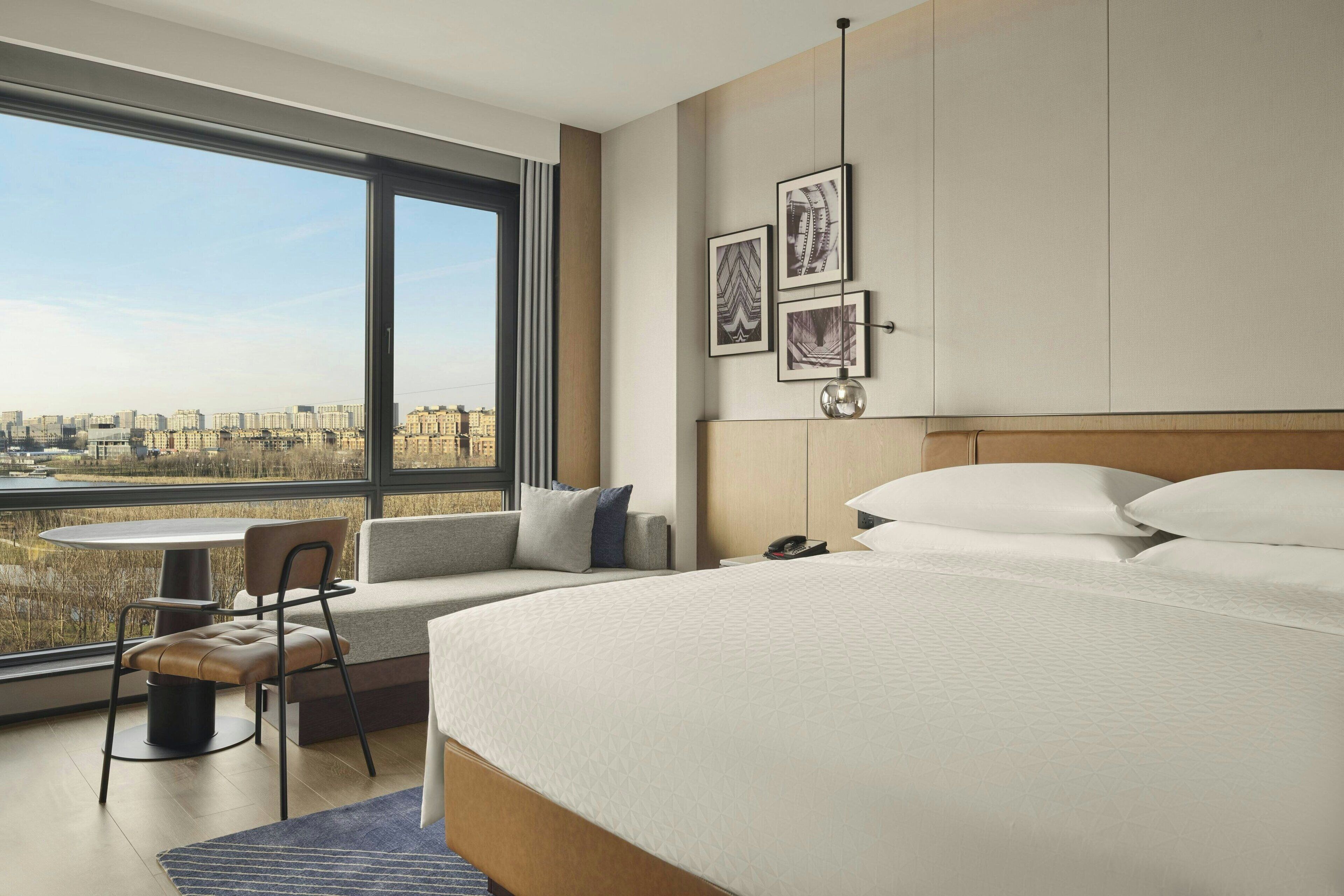 Una habitación de hotel moderna con una cama grande, una zona de estar con una mesa y vistas a la ciudad a través de amplias ventanas.
