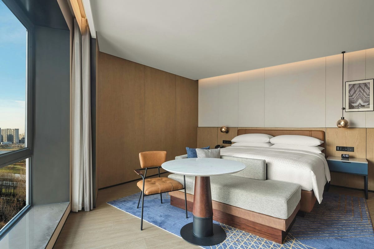 Een moderne hotelkamer met een groot bed, een ronde tafel, een stoel en uitzicht op de stad door een groot raam.