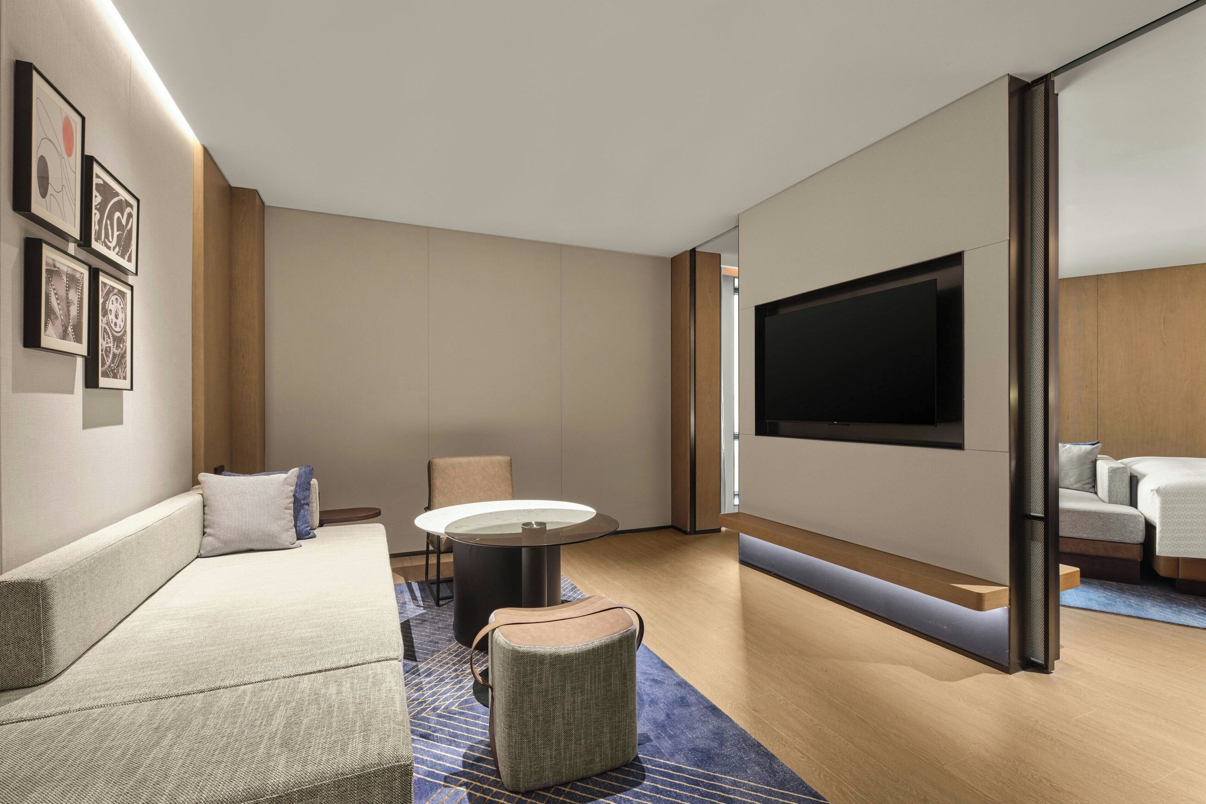 A modern hotel room with a sofa, a round table, a television mounted on the wall, and a view of the adjacent room.