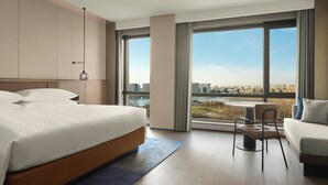 Ein modernes Hotelzimmer mit einem großen Bett, einer Couch, einem Esstisch und Blick auf eine Skyline durch große Fenster.