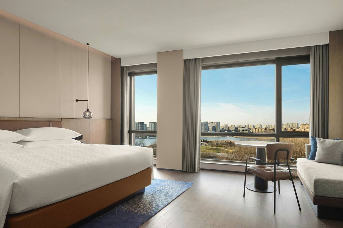 Een moderne hotelkamer met een groot bed, een bank, een eettafel en uitzicht op een stadsgezicht door grote ramen.