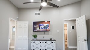 Ein Schlafzimmer mit einem Bett, einer Kommode, einem an der Wand befestigten Fernseher und zwei Türen zu anderen Räumen.