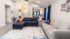 Ein Wohnzimmer mit einem Ecksofa, einem grauen Sofa und einem blauen Akzentstuhl. Man sieht einen Deckenventilator, ein Stadtbildgemälde und im Hintergrund eine Küchenzeile.