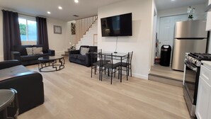 Una sala de estar moderna con un sofá, una mesa de centro y un comedor. Una escalera conduce a una cocina con un refrigerador y estufa.