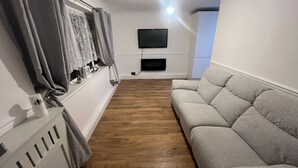 Un salon avec un canapé gris, un parquet, un téléviseur fixé au mur et une fenêtre avec des rideaux.