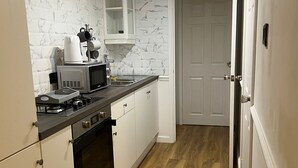 Une cuisine avec des meubles blancs, un four à micro-ondes, une cuisinière et un évier. Le sol est en bois et une porte donne accès à une autre pièce.