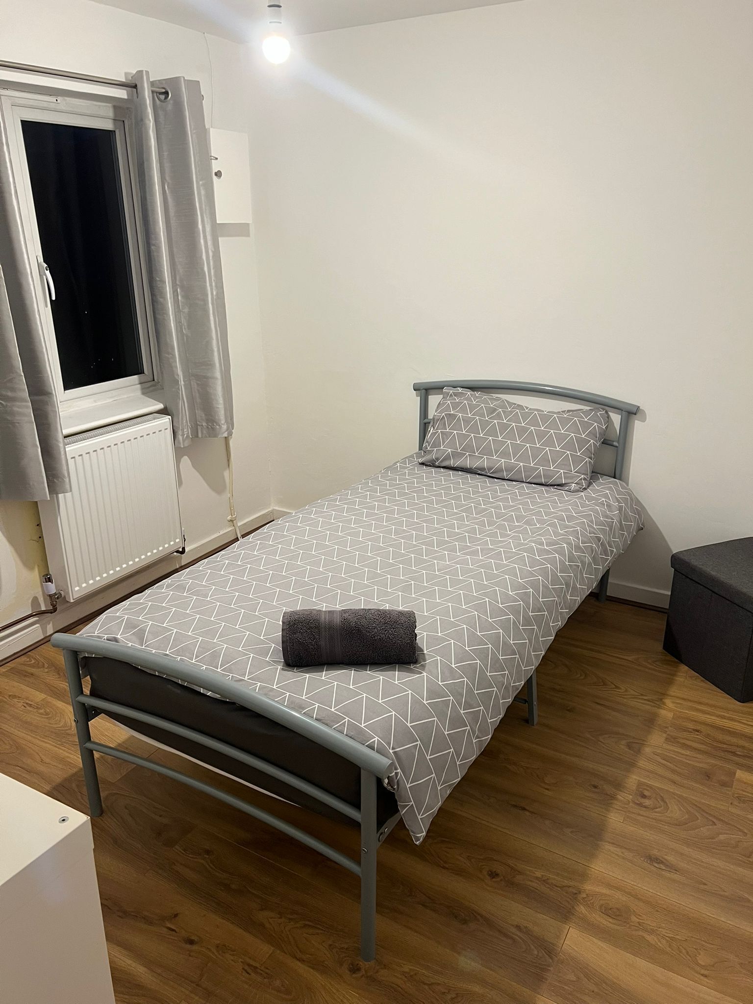 Ein Einzelbett mit grau-weißer Tagesdecke, einem grauen Kopfkissen und einer aufgerollten grauen Decke. Der Raum hat einen Holzboden, einen weißen Heizkörper und ein Fenster mit weißen Vorhängen.