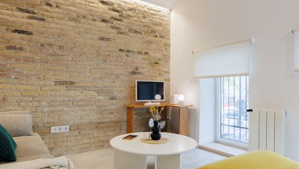 Una sala de estar moderna con una pared de ladrillo, una mesa redonda blanca con un televisor, un sillón amarillo y una ventana con vistas al exterior.