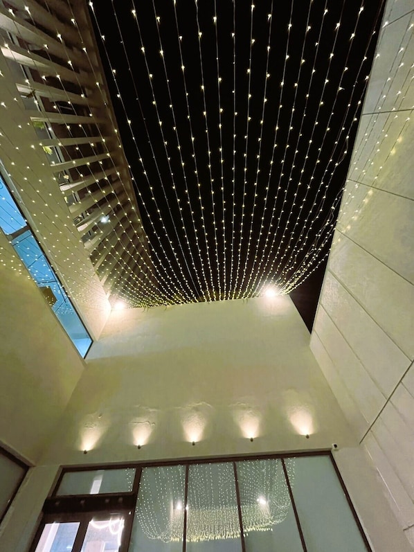 A modern interior with a high ceiling adorned with string lights, a glass wall with a view, and a clean, minimalist design. - COURT REGALIA UNIT OF S & G FESTIVITIES (Gulha)