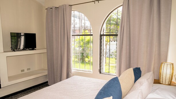 A bedroom with a bed, curtains, a television, and a window with a view of greenery.