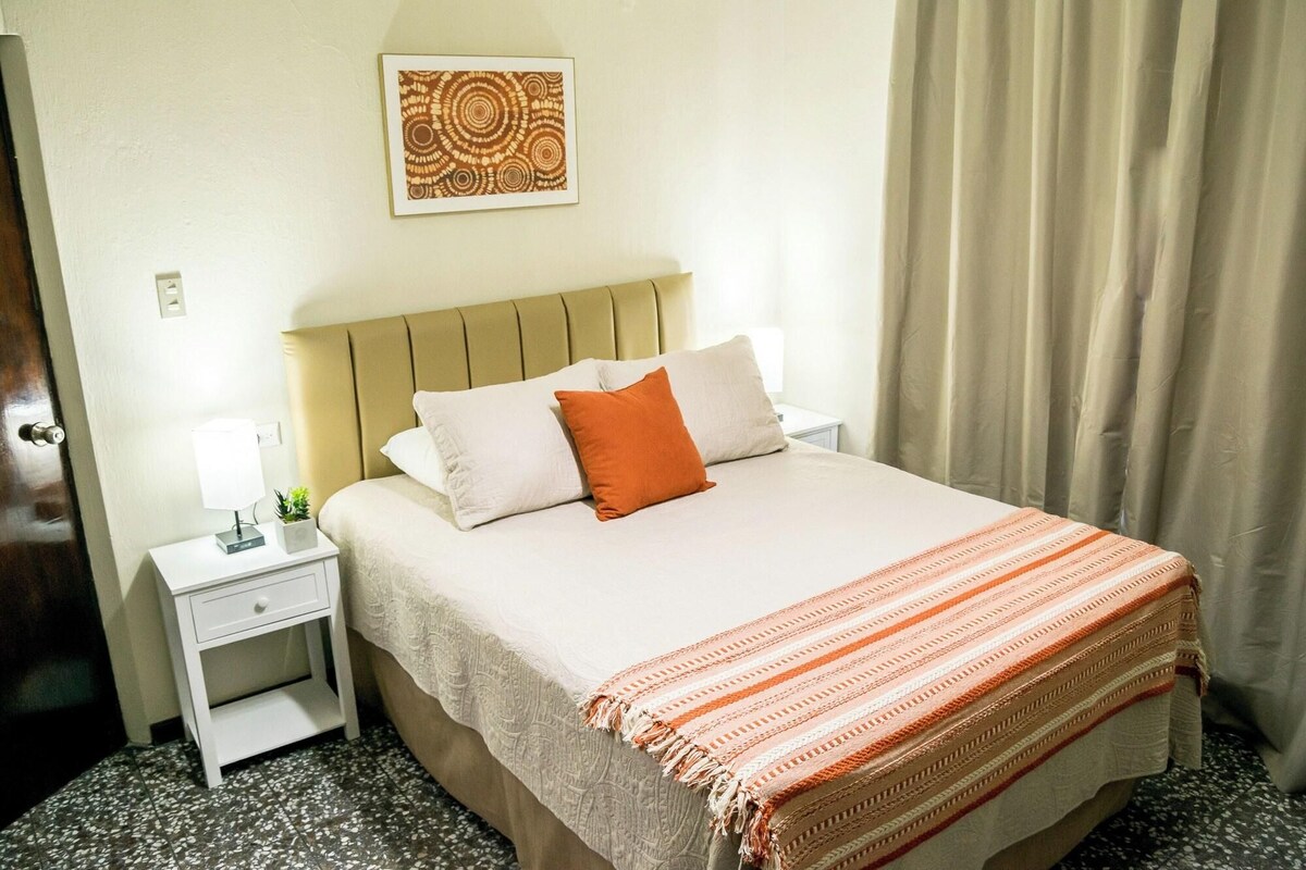Una camera d'albergo con un letto, un comodino, una lampada, una piccola pianta e un quadro appeso al muro.