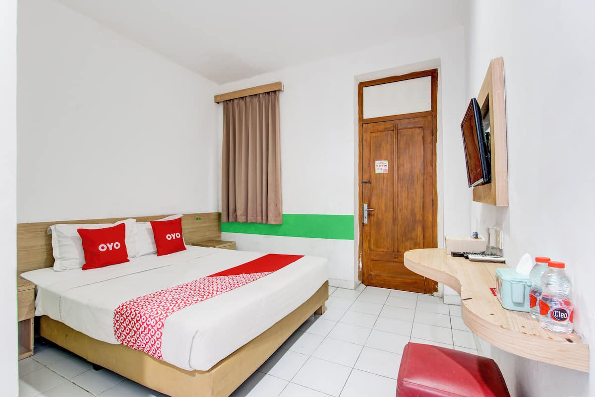 A hotel room with a bed, a wooden nightstand, a desk with a chair, a mirror, and a door.