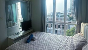 A bedroom with a bed, a TV, a window with curtains, and a city view. - NEO SEA VIEW CONDO (Pattaya)