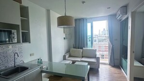 A modern kitchen with a glass-top table, a built-in oven, and a sink. There is a sofa and a view of a cityscape through a large window. - NEO SEA VIEW CONDO (Pattaya)