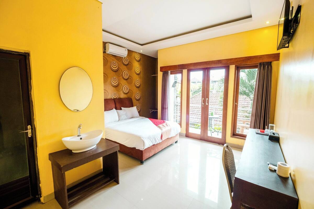 A hotel room with a bed, bedside table, and a desk with a chair. There is a window with a view of greenery outside.