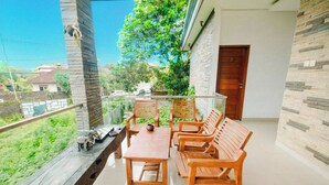 A balcony with a wooden table and chairs, a glass railing, and a view of greenery and houses. - Aeena Homestay (Ungasan)