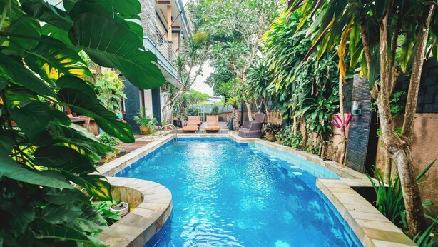 A rectangular swimming pool with a tiled edge, surrounded by lush greenery and a few lounge chairs. - Aeena Homestay (Ungasan)