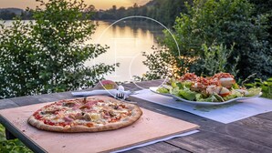 A wooden table with a pizza and a salad plate, set against a backdrop of a lake and trees during sunset.