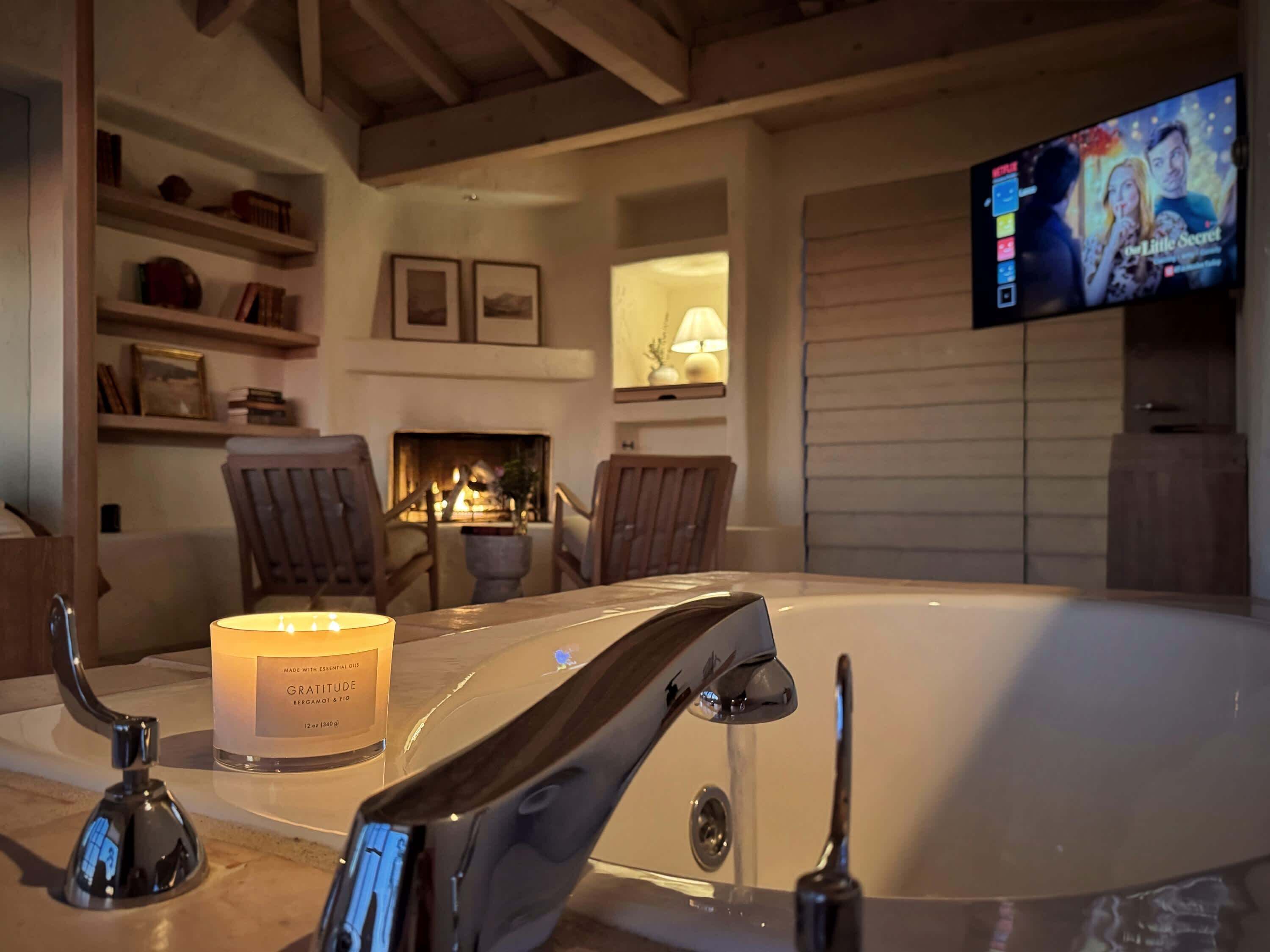 A modern bathroom with a bathtub, a lit candle, and a television displaying a movie.