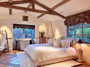 A bedroom with a large bed, a desk, and a window with a view of greenery.