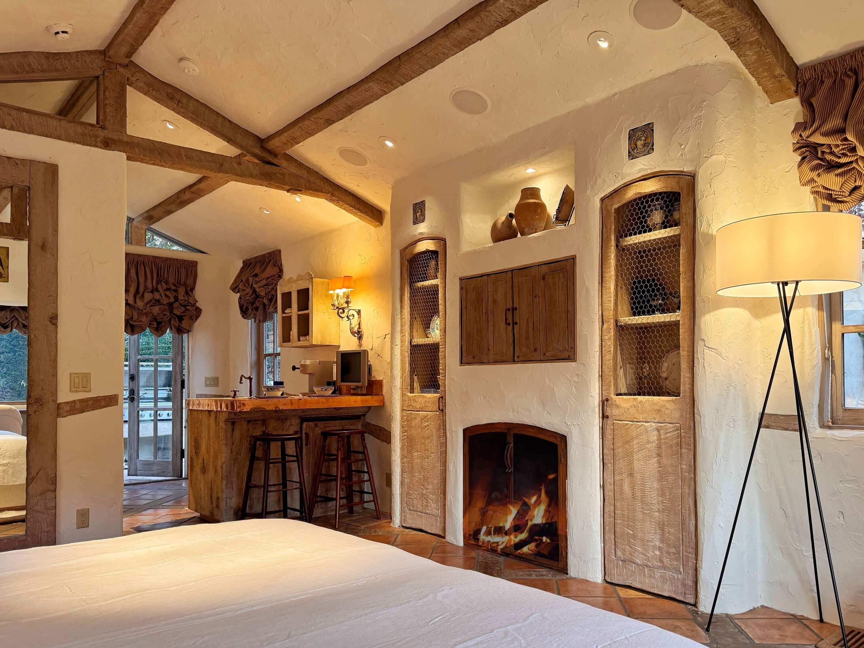 A spacious bedroom with a large bed, a fireplace, and a kitchen area visible through an open doorway.